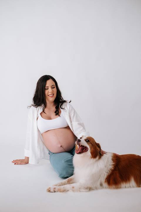Foto da gestante sentada no chão sorrindo para o cão de estimação.'
