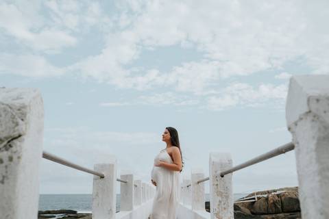 Foto da gestante de vestido branco em uma passarela sobre as rochas.'