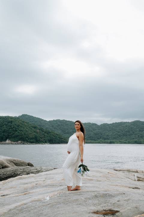 Foto da gestante de perfil com montanhas e mar ao fundo.'