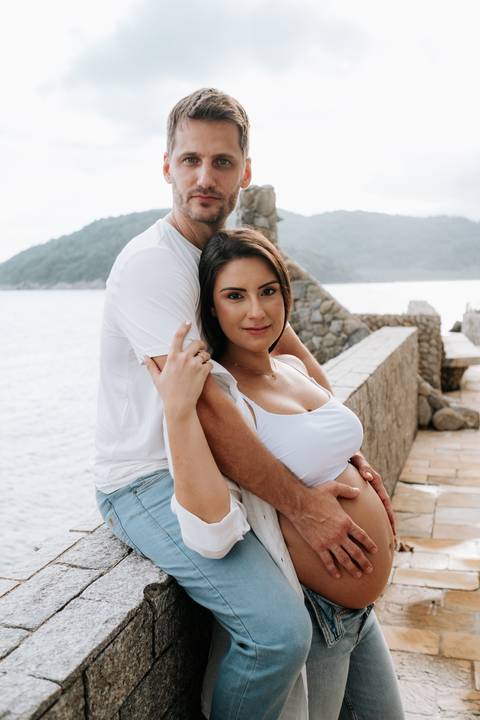 Foto da gestante sorrindo enquanto é abraçada por trás pelo parceiro.'
