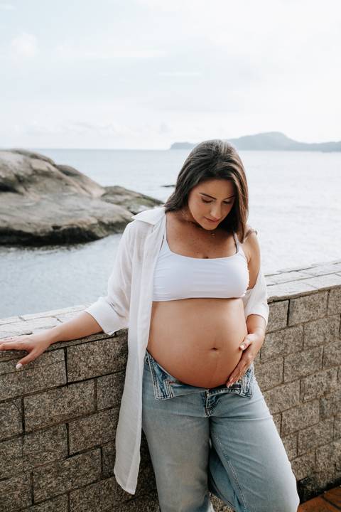 Foto da gestante de top branco destacando a barriga em frente ao mar.'