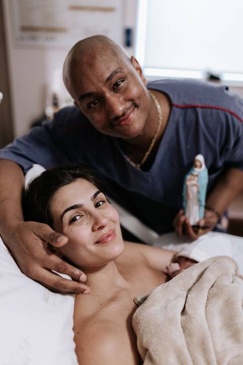 Foto do casal sorrindo com a bebê e uma imagem religiosa.'