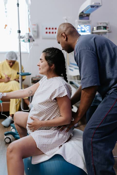 Foto da gestante sentada na bola de pilates com apoio do parceiro.'