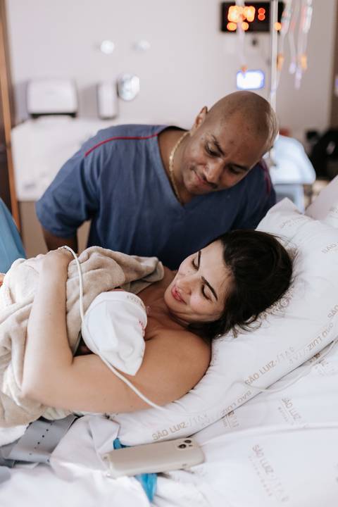 Foto do casal admirando a bebê recém-nascida na cama hospitalar.'
