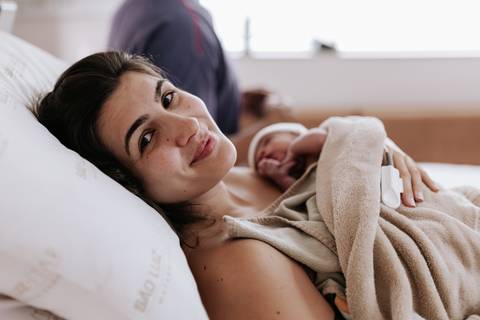 Foto da mãe sorrindo com a bebê enrolada em uma manta.'