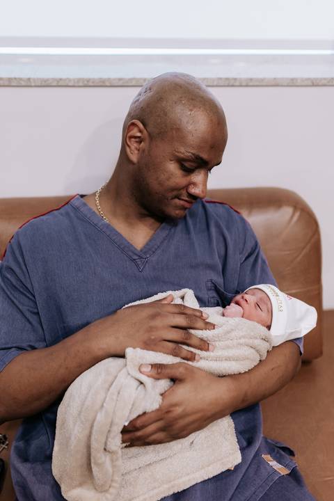 Foto do pai segurando a bebê com ternura na poltrona da maternidade.'