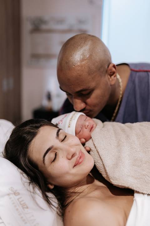 Foto do pai beijando a cabeça da bebê com a mãe ao lado.'