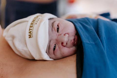 Foto da bebê em contato pele a pele com a mãe sob manta azul.'