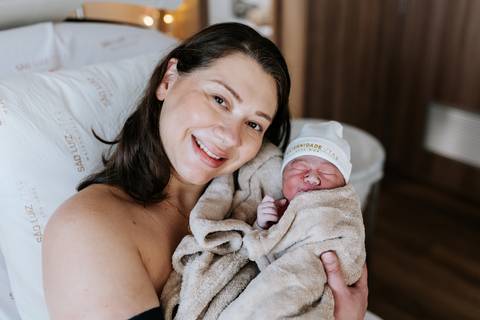 Foto da mãe sorridente segurando a bebê envolvida em uma manta.'