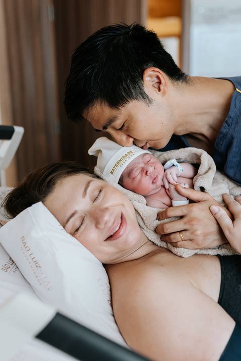 Foto do pai beijando a testa da bebê com a mãe do lado.'