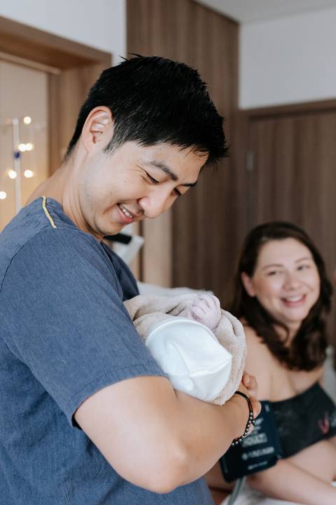 Foto do pai admirando a bebê enquanto a mãe sorri ao fundo.'