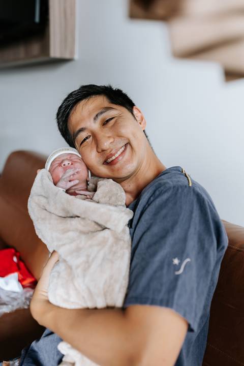 Foto do pai abraçado à bebê recém-nascida sob luz suave.'