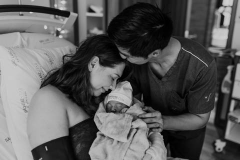 Foto em preto e branco do casal abraçado com a bebê recém-nascida.'