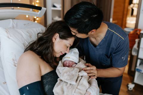 Foto do pai beijando a cabeça da mãe enquanto ela segura a bebê.'