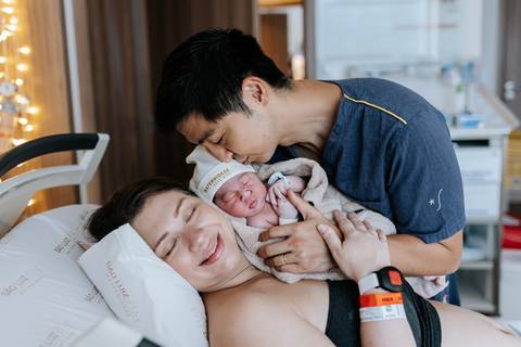 Foto do pai beijando a cabeça da mãe com a bebê no colo na maternidade.'