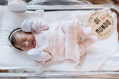 Foto da bebê no berço hospitalar com saída de maternidade rosa.'