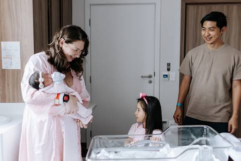 Foto da família reunida no quarto com a irmã mais velha conhecendo a bebê.'
