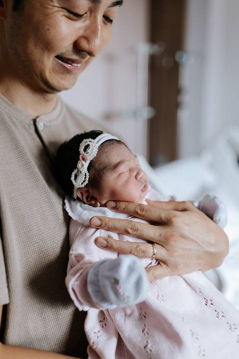 Foto da mão do pai amparando a bebê no colo em plano detalhe.'