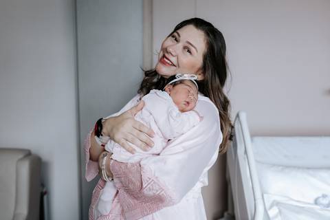 Foto da mãe em pé segurando a bebê no quarto da maternidade.'