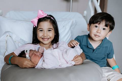 Foto dos dois irmãos mais velhos conhecendo a bebê na cama.'