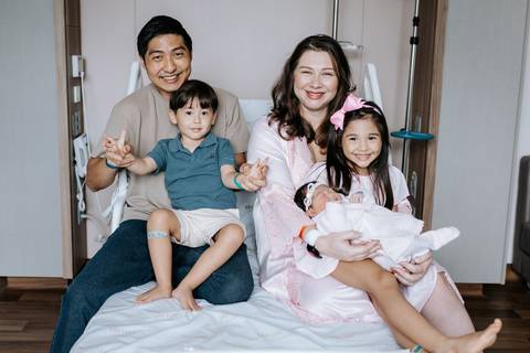 Foto da família completa reunida na cama da maternidade sorrindo.'
