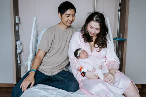 Foto da mãe com a bebê no colo ao lado do pai.'