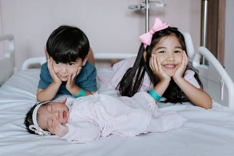 Foto dos dois irmãos mais velhos deitados ao lado da bebê sorrindo.'