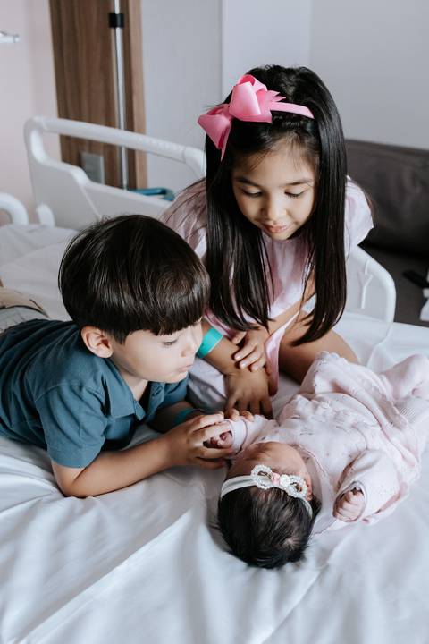Foto dos irmãos observando e tocando a bebê com curiosidade e afeto.'