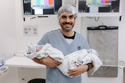 Foto do pai orgulhoso segurando os dois filhos no colo no centro cirúrgico.'