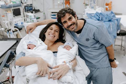 Foto do casal sorridente abraçado com os dois bebês no pós-parto.'
