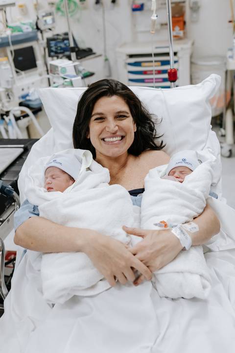 Foto da mãe radiante segurando os gêmeos no colo na sala de recuperação.'
