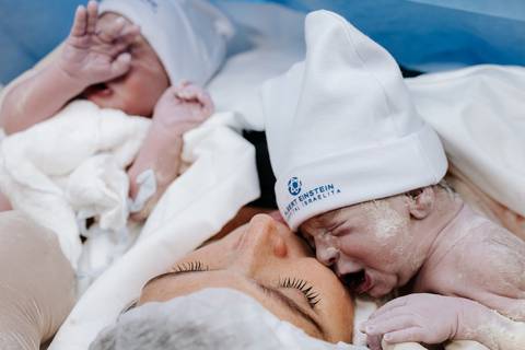 Foto da mãe de olhos fechados sentindo os dois bebês próximos ao rosto.'