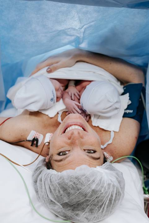 Foto da mãe radiante sorrindo com os dois bebês sobre o peito.'