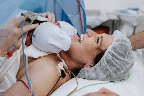 Foto da mãe rindo de emoção com os bebês no pós-parto imediato.'