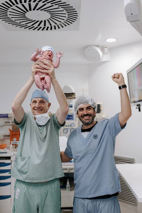 Foto do pai e do médico com o bebê vibrando e comemorando o nascimento.'