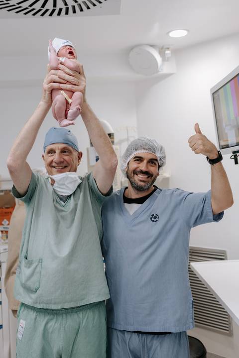 Foto do pai sorridente fazendo sinal de positivo com o médico e o bebê.'