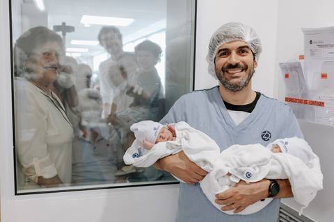 Foto do pai apresentando os gêmeos para a família pelo visor do berçário.'