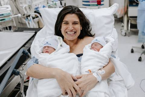 Foto da mãe radiante segurando os gêmeos no colo na sala de recuperação.'