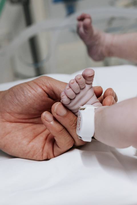 Foto em detalhe da mão do pai segurando o pé da recém-nascida.'