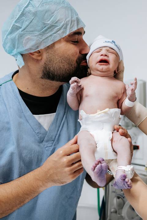 Foto do pai beijando o bebê durante a avaliação neonatal.'