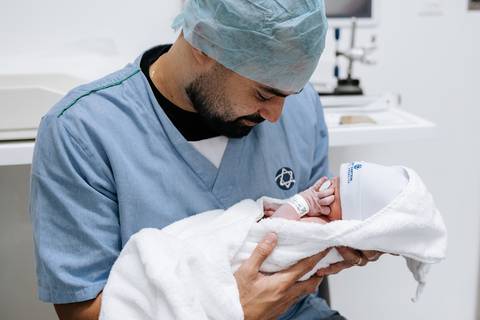 Foto do pai segurando o bebê envolvida em manta branca pela primeira vez.'