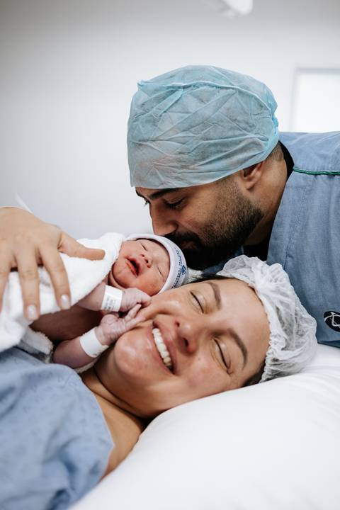Foto do pai beijando a cabeça da mãe enquanto ela segura o bebê.'