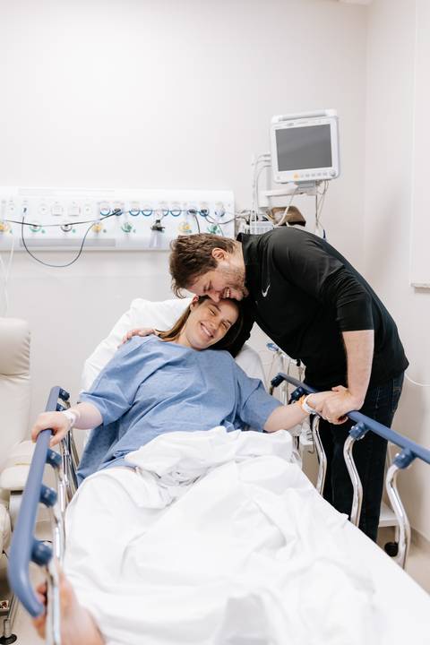 Foto do casal sorrindo na maca hospitalar antes do parto.'