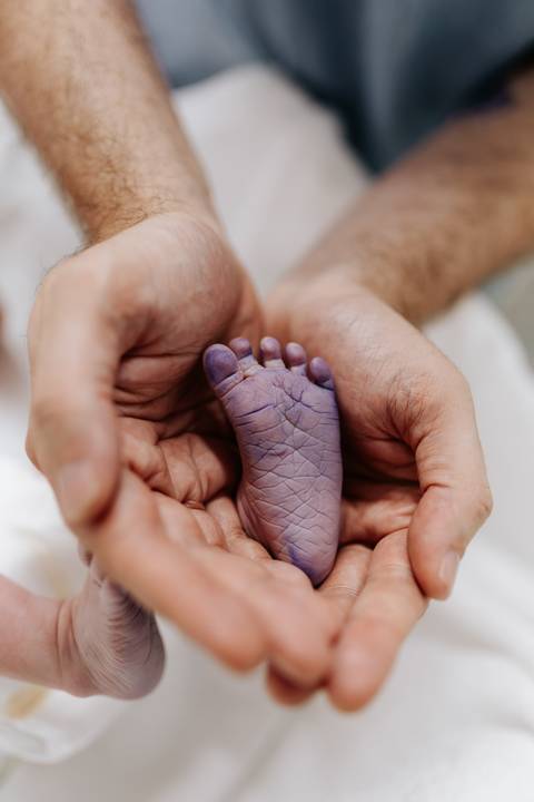 Mãos do pai em concha segurando o pé do recém-nascido.'