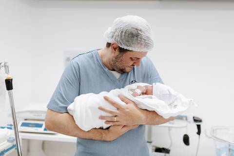 Pai segurando o filho no colo, envolto em manta branca.'