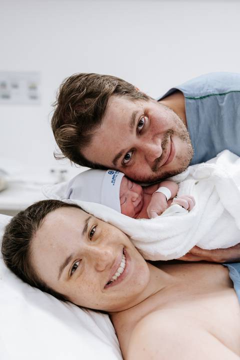 Casal sorridente com o bebê posicionado entre seus rostos.'
