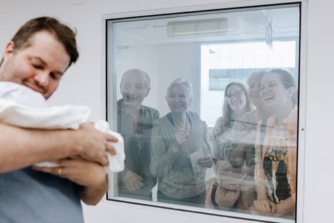 Pai apresentando o bebê para a família através do vidro do berçário.'