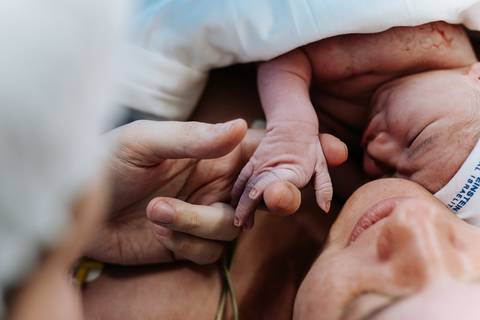 Detalhe das mãos do bebê segurando o dedo da mãe no contato pele a pele.'