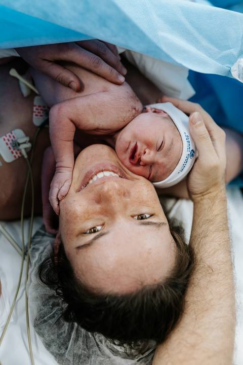 Close da mãe sorrindo com o bebê de touca azul em seu peito.'