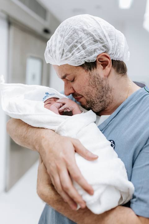 Pai segurando o filho no colo, envolto em manta branca, com olhar sereno.'